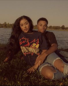 two people sitting next to each other on the grass near a body of water with trees in the background