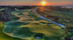 an aerial view of a golf course with the sun setting over the water in the background