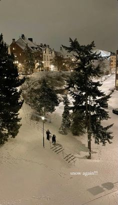 two people walking up the side of a snow covered ski slope at night with buildings in the background