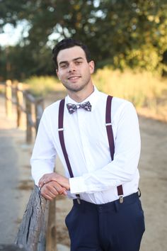 a man in a white shirt and bow tie leaning on a fence with his arms crossed