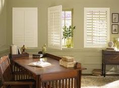 a wooden desk sitting in front of two windows with shutters on each side and a potted plant next to it