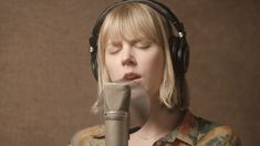 a woman wearing headphones and singing into a microphone with her eyes closed, in front of a brown background