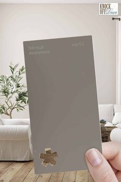 a person holding up a gray paint swatch in front of a living room with white furniture