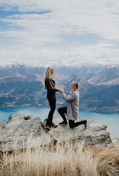 two women sitting on top of a large rock near the ocean and mountains holding hands