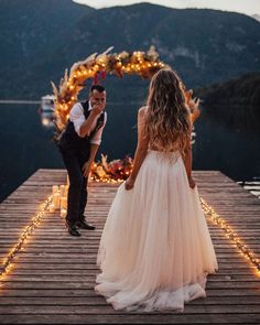 a bride and groom standing on a dock with candles in front of them, looking at each other