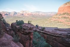 two people are standing on the edge of a cliff with trees and mountains in the background