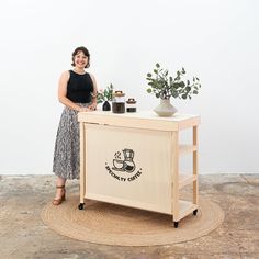 a woman standing next to a table with plants and jars on it in front of a white wall