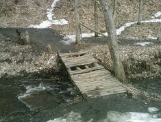 a small wooden bridge over a stream in the woods with snow on the ground and trees