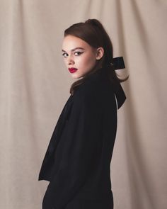 a woman with long hair wearing a black jacket and red lipstick standing in front of a white backdrop