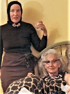 two women sitting on a bed with one holding a wine glass and the other looking at the camera