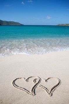 two hearts drawn in the sand on a beach with blue water and mountains in the background