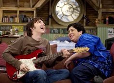 two young men sitting on a couch playing guitars and singing into each other's mouths