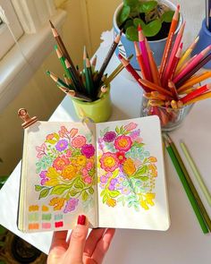 a person holding an open notebook with colored pencils in front of them on a table