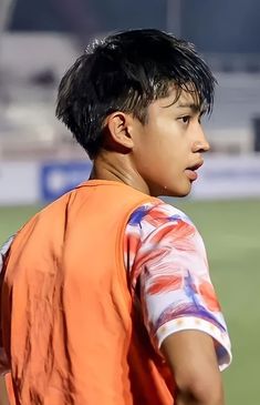 a young boy standing on top of a soccer field wearing an orange shirt and black hair