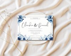 a white and blue wedding card on top of a bed next to some dried flowers