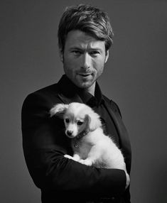 a man holding a small white dog in his arms and looking at the camera while wearing a black shirt