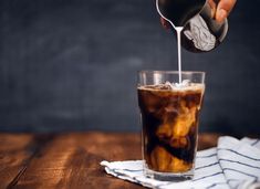 a person pouring ice into a glass filled with iced coffee on top of a wooden table