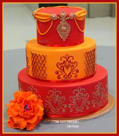 a three tiered cake decorated with red, yellow and orange designs on a table next to an orange flower