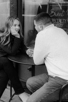 a man and woman sitting at a table in front of a store window, talking to each other