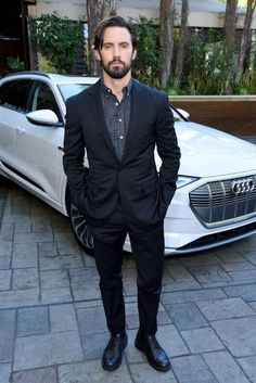 a man standing in front of a car wearing a black suit and tie with his hands in his pockets