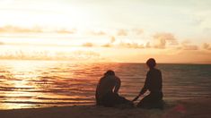 two people are sitting on the beach watching the sun go down over the water and clouds in the sky