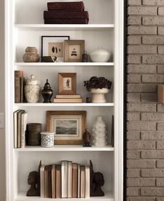 a white book shelf filled with lots of books next to a brick wall in a living room