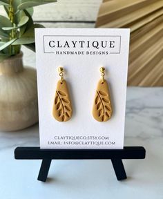 a pair of gold earrings with leaves on them sitting next to a card that says claytiquee handmade designs