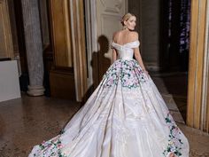 a woman in a white dress with flowers on the skirt is standing near columns and looking off to the side