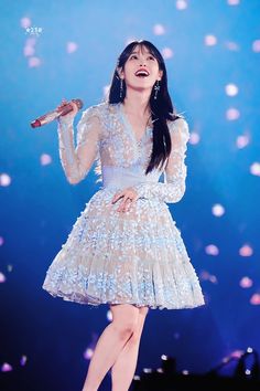 a woman in a short white dress holding a microphone and singing on stage with lights behind her