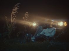 two people sitting in front of a car at night with headlights shining on the ground