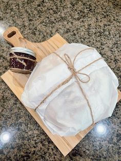 a wooden cutting board topped with a jar of jam next to a wrapped bag on top of a counter