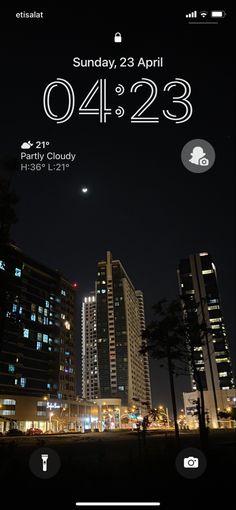 an iphone screen showing the date and time in front of some tall buildings at night