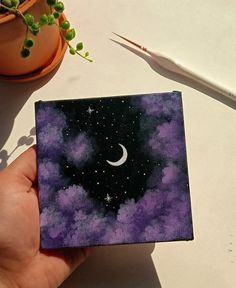 a hand holding a small square piece of paper with the moon and stars painted on it