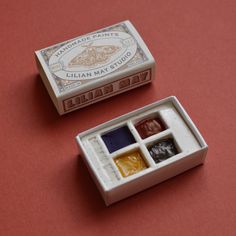 two miniature boxes with different colored paints in them on a red table top next to each other