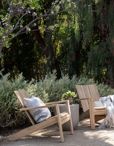 two wooden chairs sitting next to each other on a dirt ground near trees and bushes