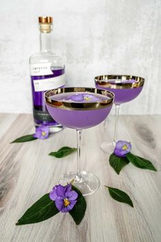 two martini glasses with purple flowers on the rims and one glass is filled with liquid