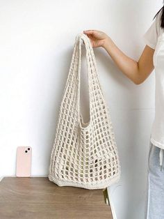 a woman holding a crocheted white bag next to a phone on a table