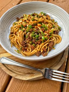 a white bowl filled with pasta and meat on top of a wooden table next to a fork