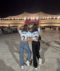 two young women standing in front of a building at night with their arms around each other