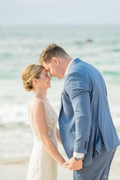 a bride and groom standing on the beach holding each other's hands while looking at each other