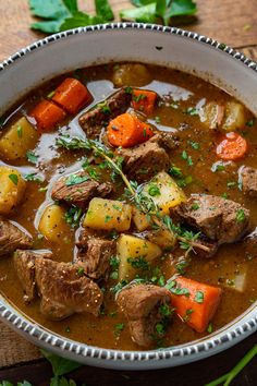 a bowl filled with stew and carrots on top of a wooden table next to parsley