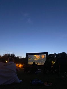 people sitting in the grass watching a movie on a large screen at night with bright lights