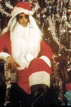 a man dressed as santa claus sitting in front of a christmas tree holding a bottle