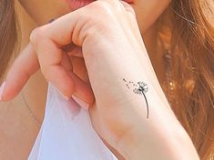 a woman with a small tattoo on her left wrist and hand is holding a dandelion in front of her face