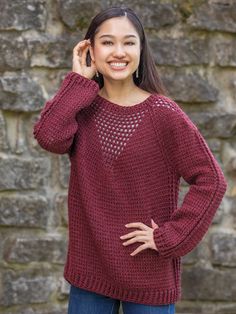 a woman wearing a red sweater and jeans standing in front of a stone wall with her hands on her head