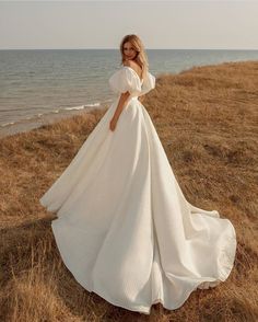 a woman standing on top of a grass covered field next to the ocean wearing a white dress