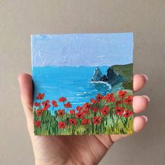 a hand holding up a small piece of art with red flowers on the grass and water in the background