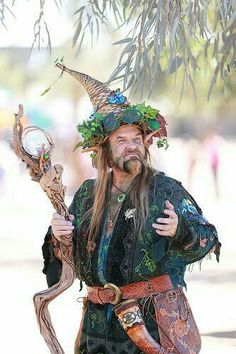 a man dressed in costume holding a clock and wearing a horned headdress while standing under a tree