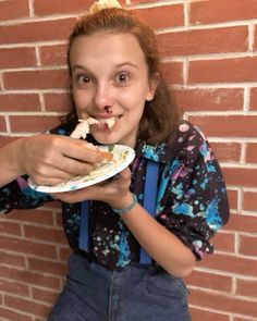 a girl eating food from a paper plate in front of a brick wall with her hands