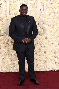 a man in a tuxedo standing on a red carpet with flowers behind him